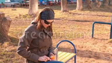 年轻的黑发女人戴着太阳镜，<strong>剥</strong>着<strong>橘子</strong>坐在公园的长凳上。